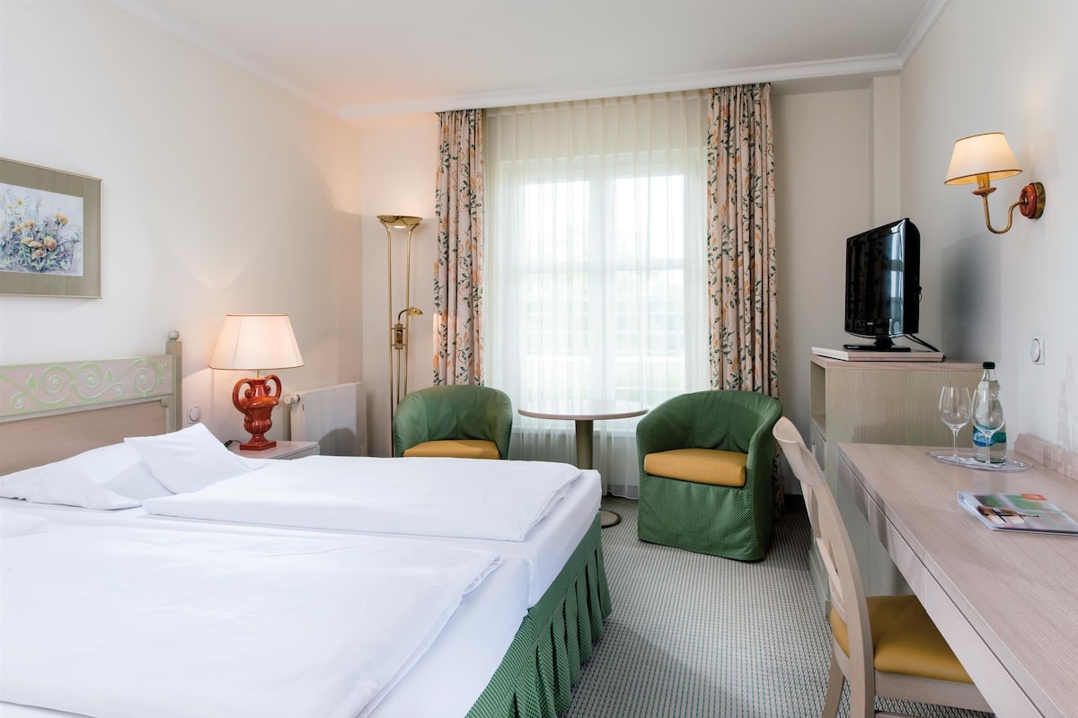 A hotel room with a bed, two chairs, a desk with a television, a lamp, and a window with curtains.