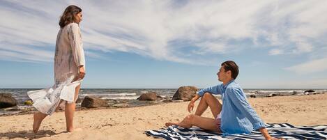 A beach scene with a person sitting on a striped beach towel and another person walking.