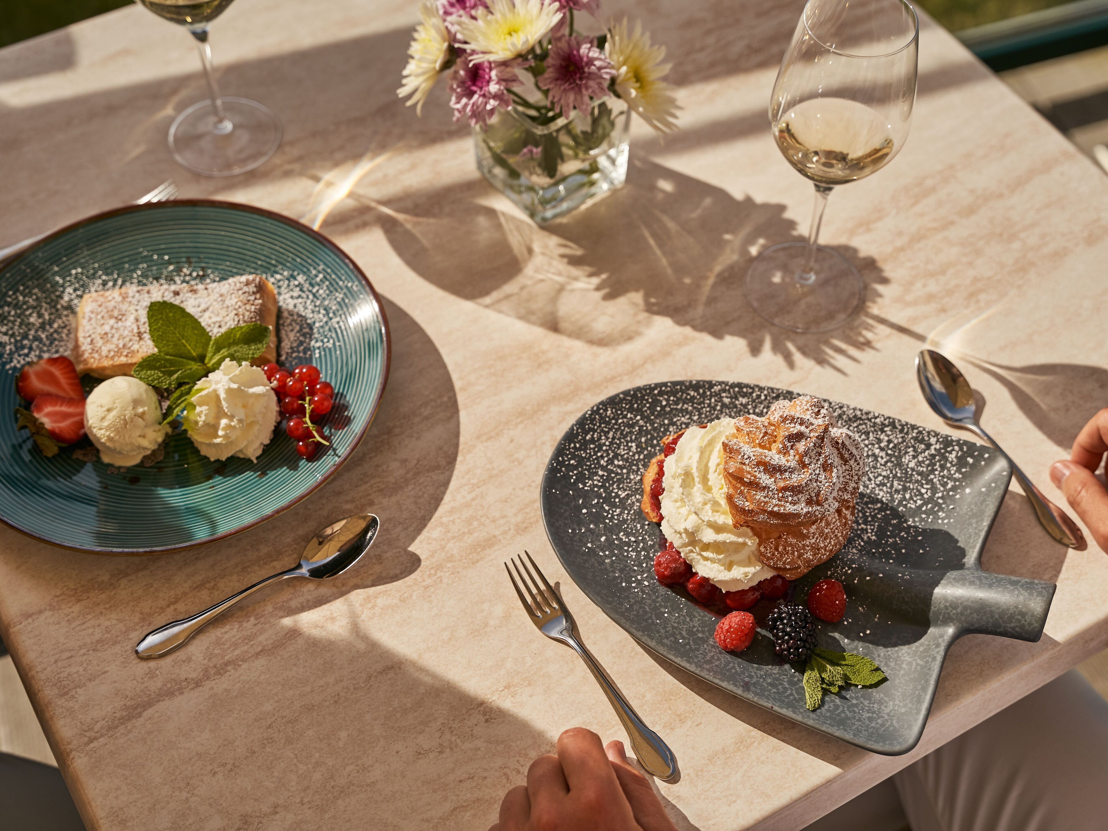 A table with two plates of dessert, a vase with flowers, and a glass of wine.