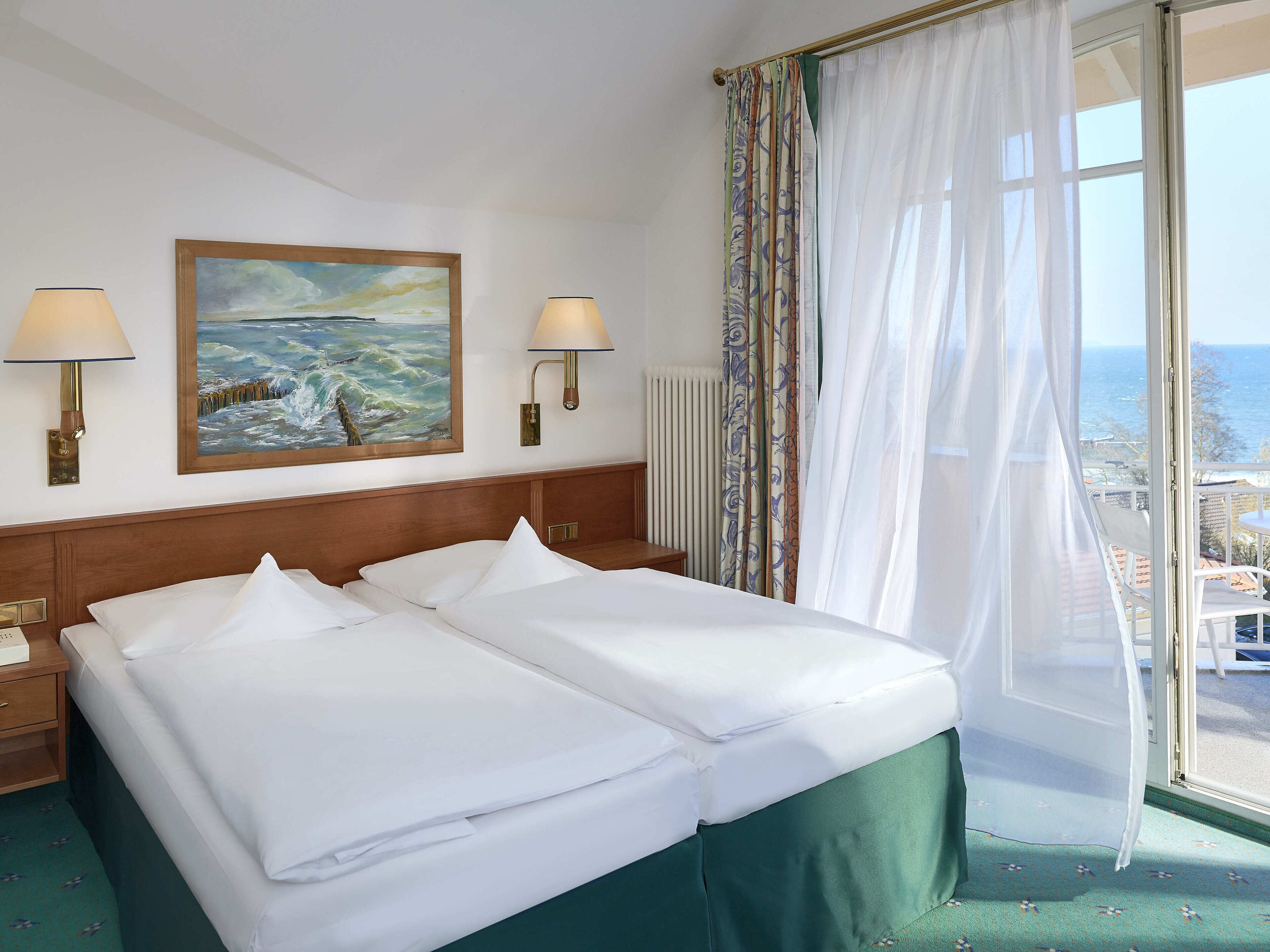 A hotel room with two beds, a painting on the wall, two wall lamps, and a view of the sea through sheer curtains.
