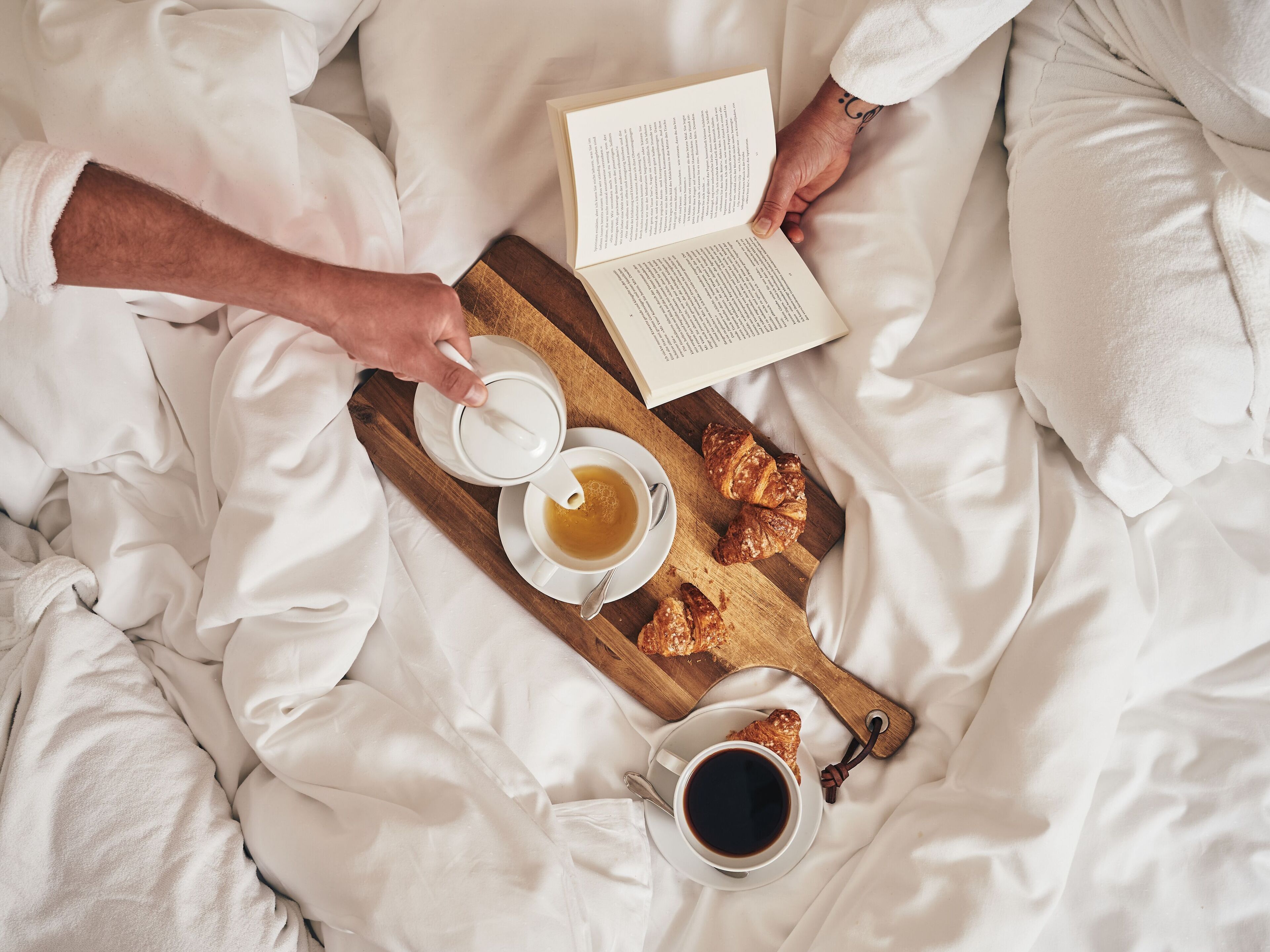 A person lying in bed with a book, a cup of tea, and croissants on a wooden tray.
