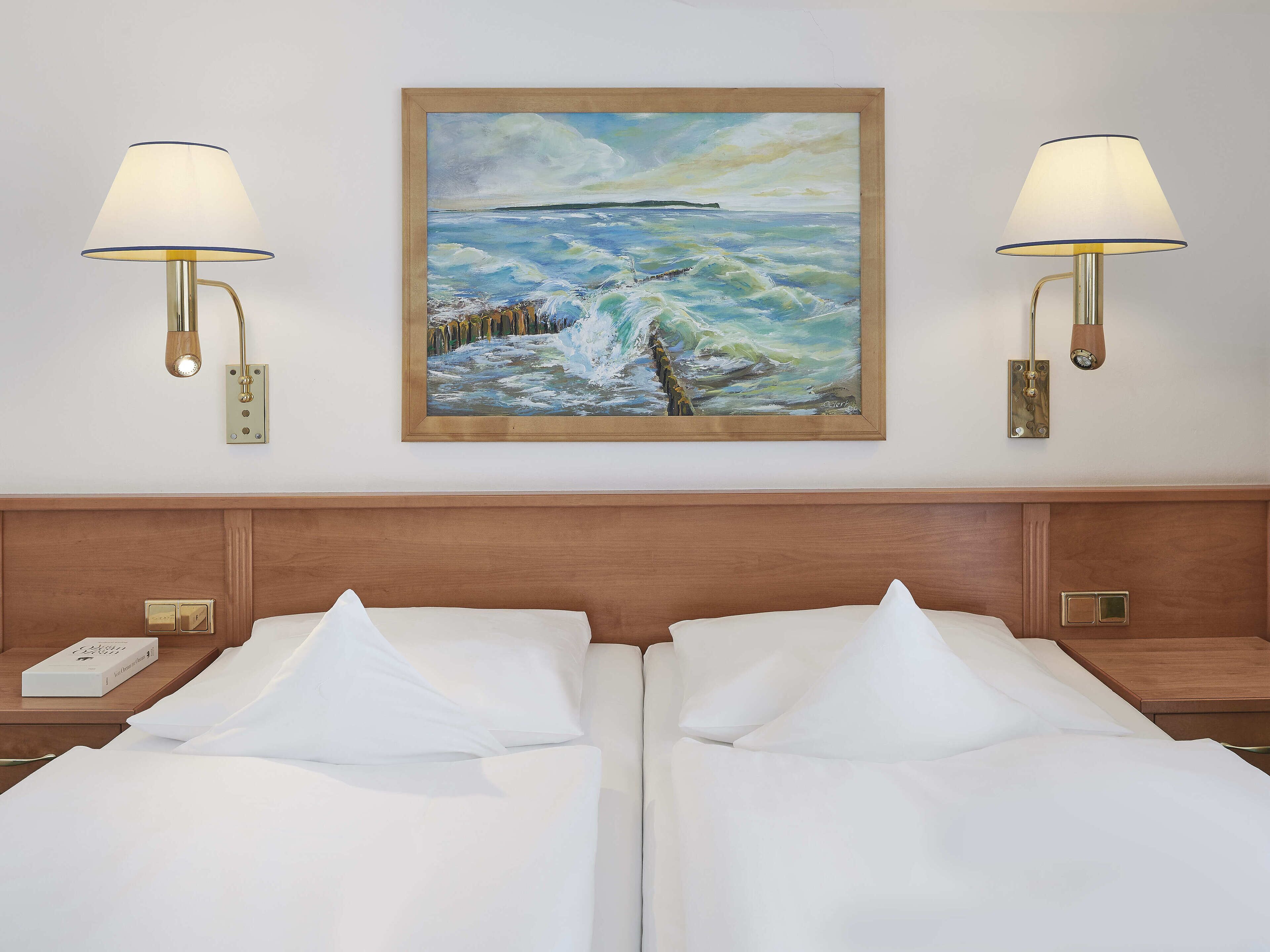 A hotel room with two beds, a painting of a seascape, and two wall-mounted lamps.