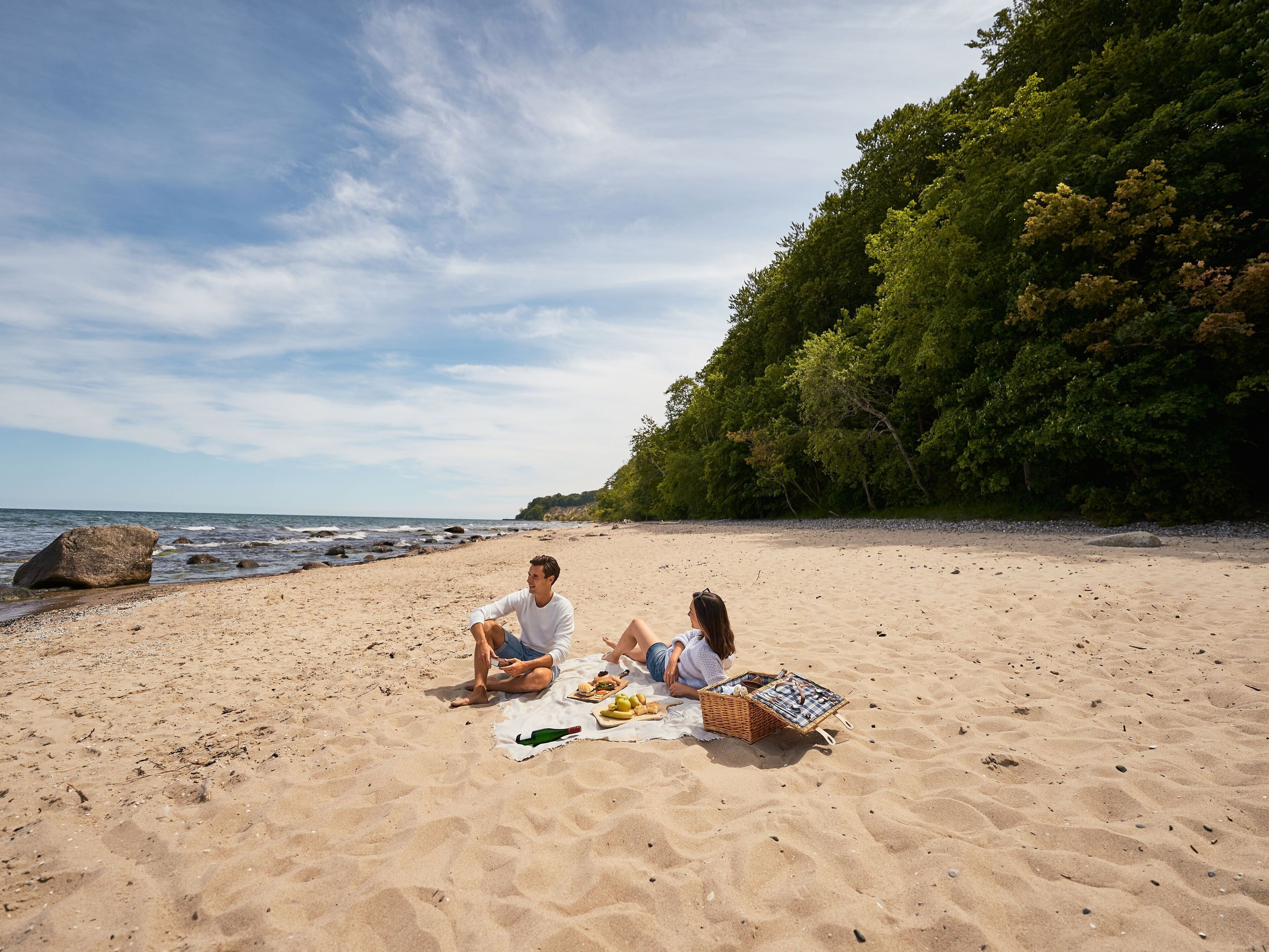 A beach with two people sitting on a blanket, a picnic basket, and a bottle.