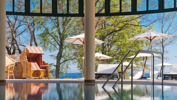 An indoor swimming pool with clear views of the sea through large windows, surrounded by lounge chairs and umbrellas.
