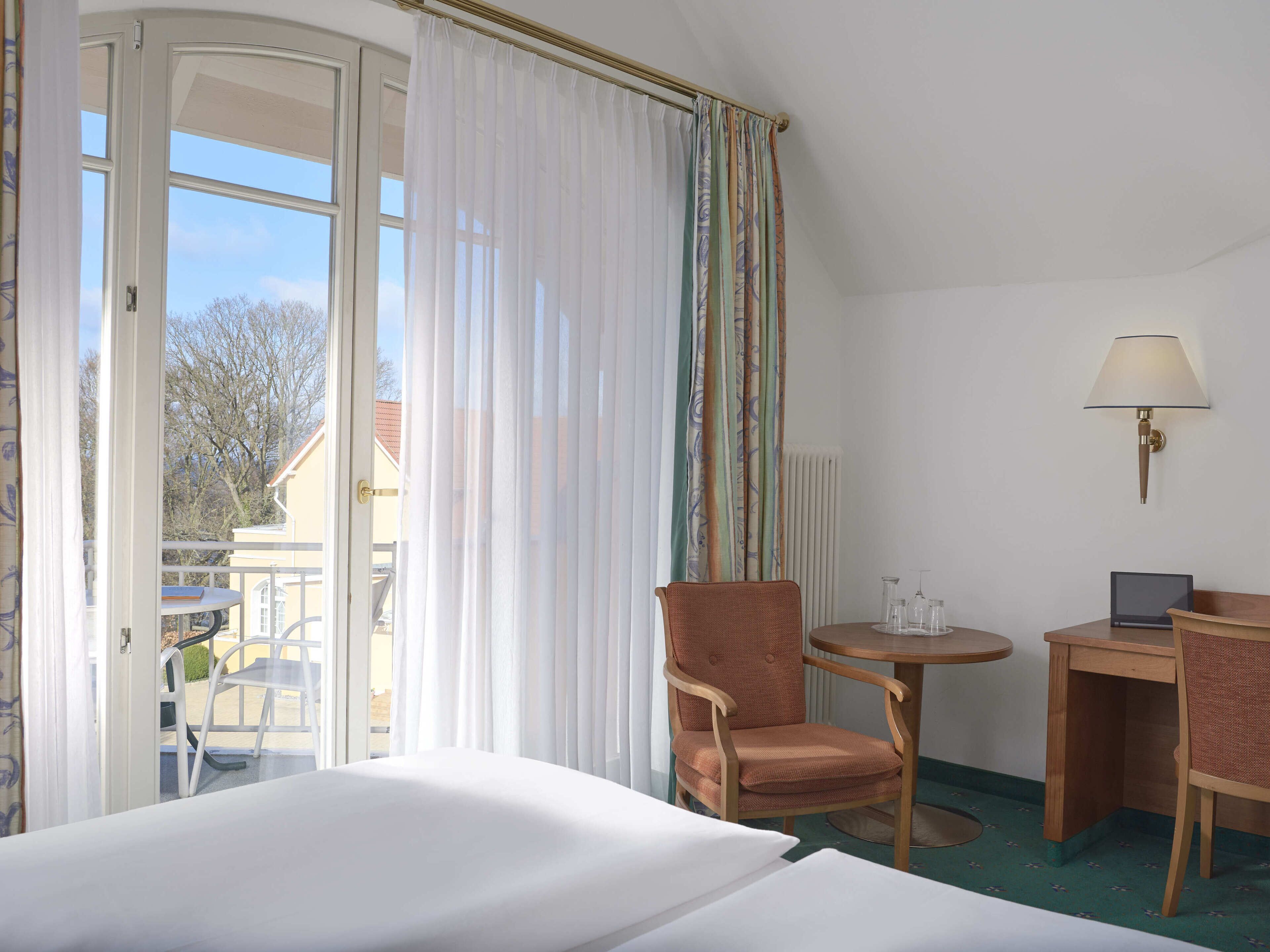 Ein Hotelzimmer mit Bett, Sessel, Nachttisch, Lampe und Blick nach draußen.