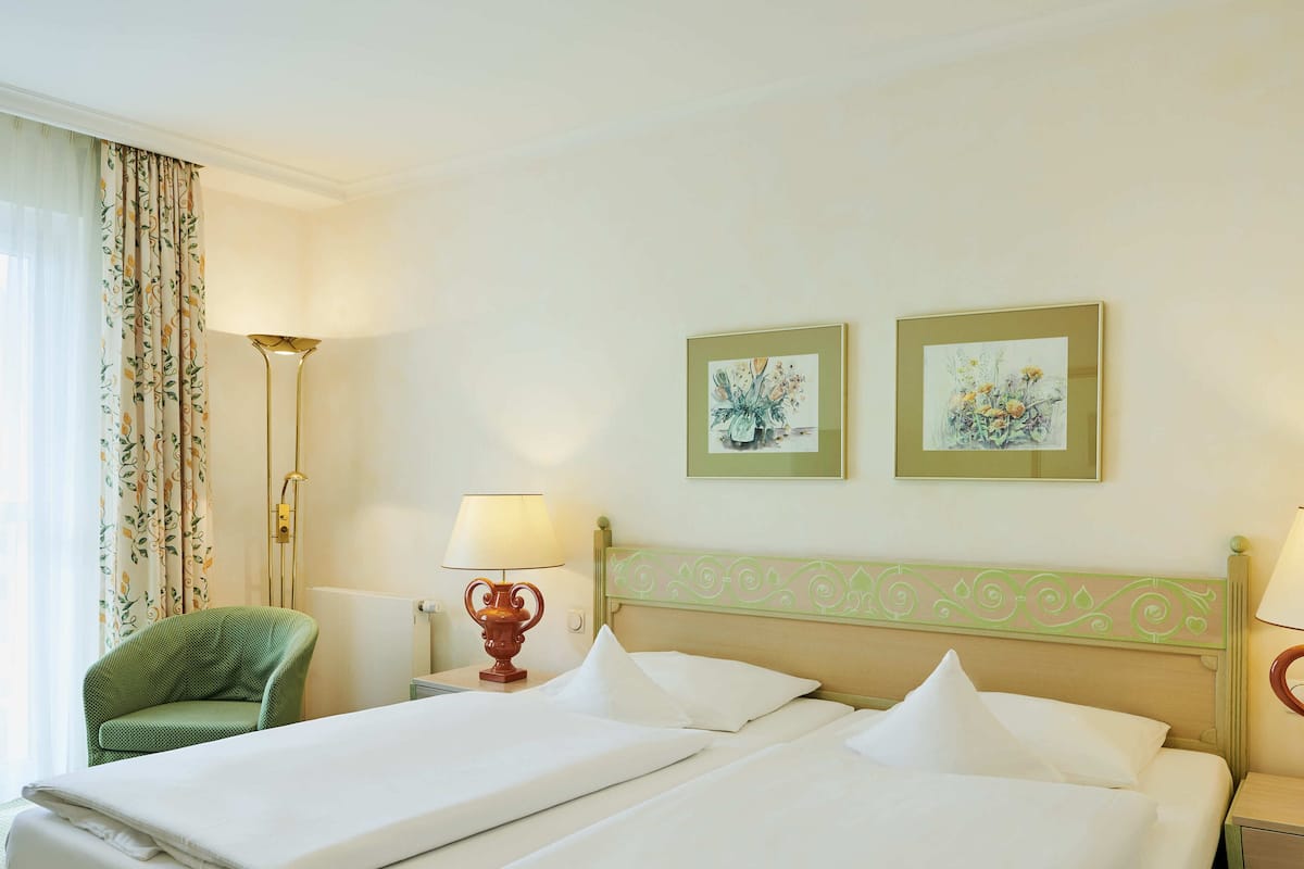 A hotel room with two beds, a green armchair, a floor lamp, and two framed floral paintings on the wall.