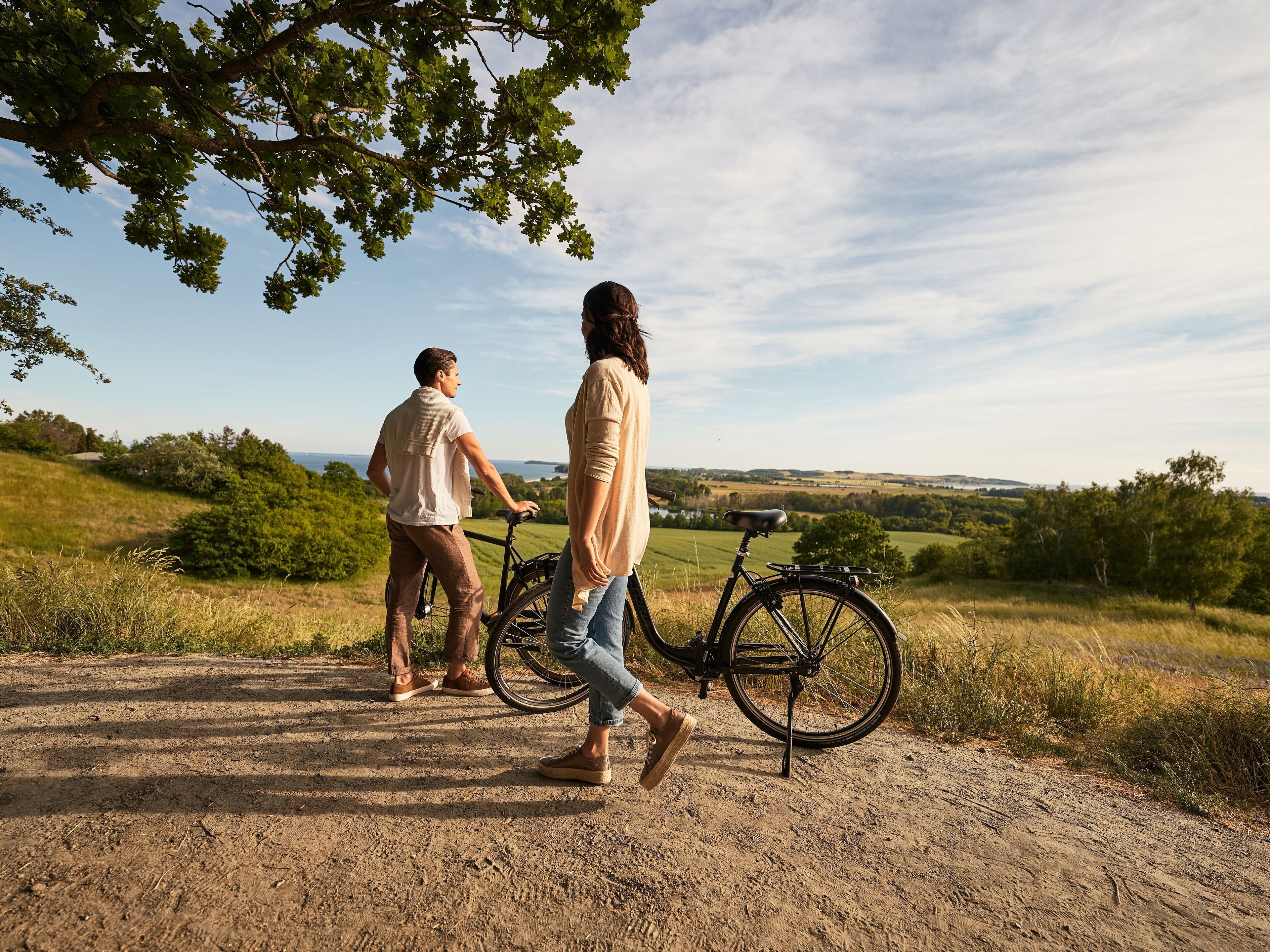 Två personer promenerar på en grusväg med en cykel, omgivna av grönska och en klar himmel.
