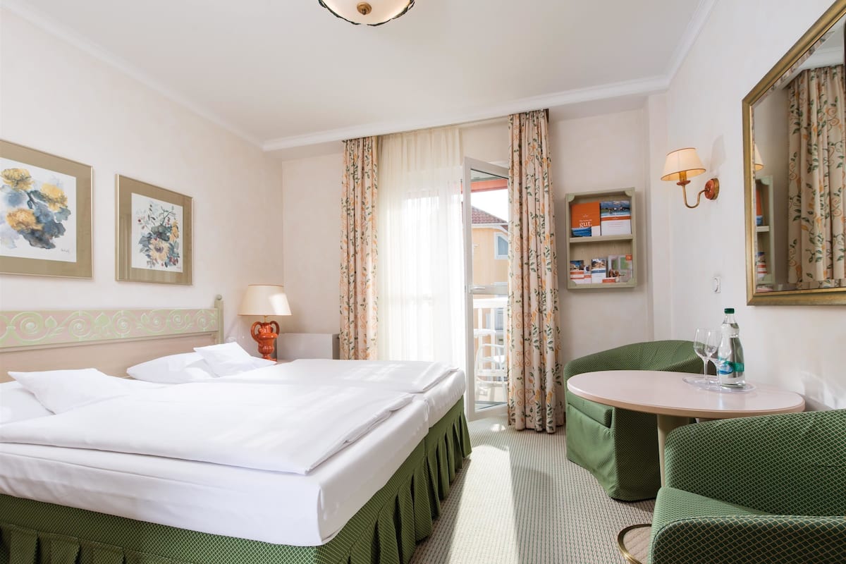 A hotel room with a bed, a small table with a chair, a lamp, and a bookshelf.