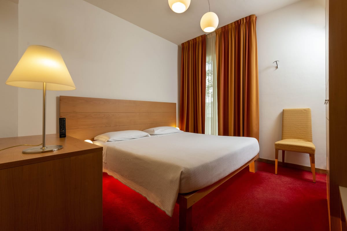 A hotel room with a wooden headboard, a bed with white linens, a red carpet, a lamp, a chair, and a window with curtains.