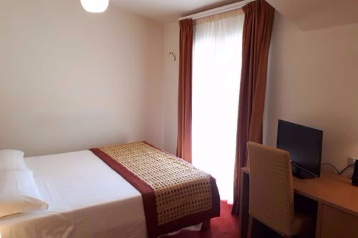 A hotel room with a bed, a desk with a computer, and a window with curtains.