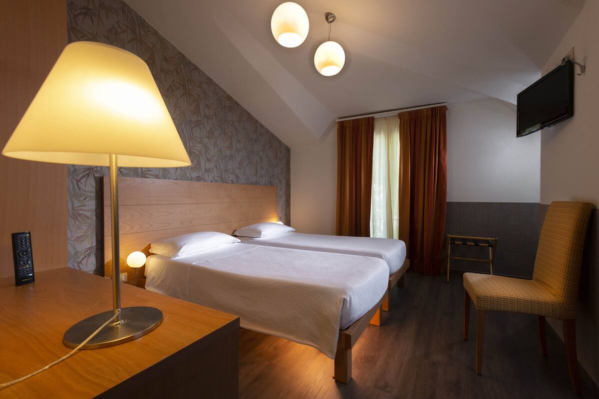 A hotel room with two single beds, a desk with a lamp, a chair, a TV mounted on the wall, and a small table.