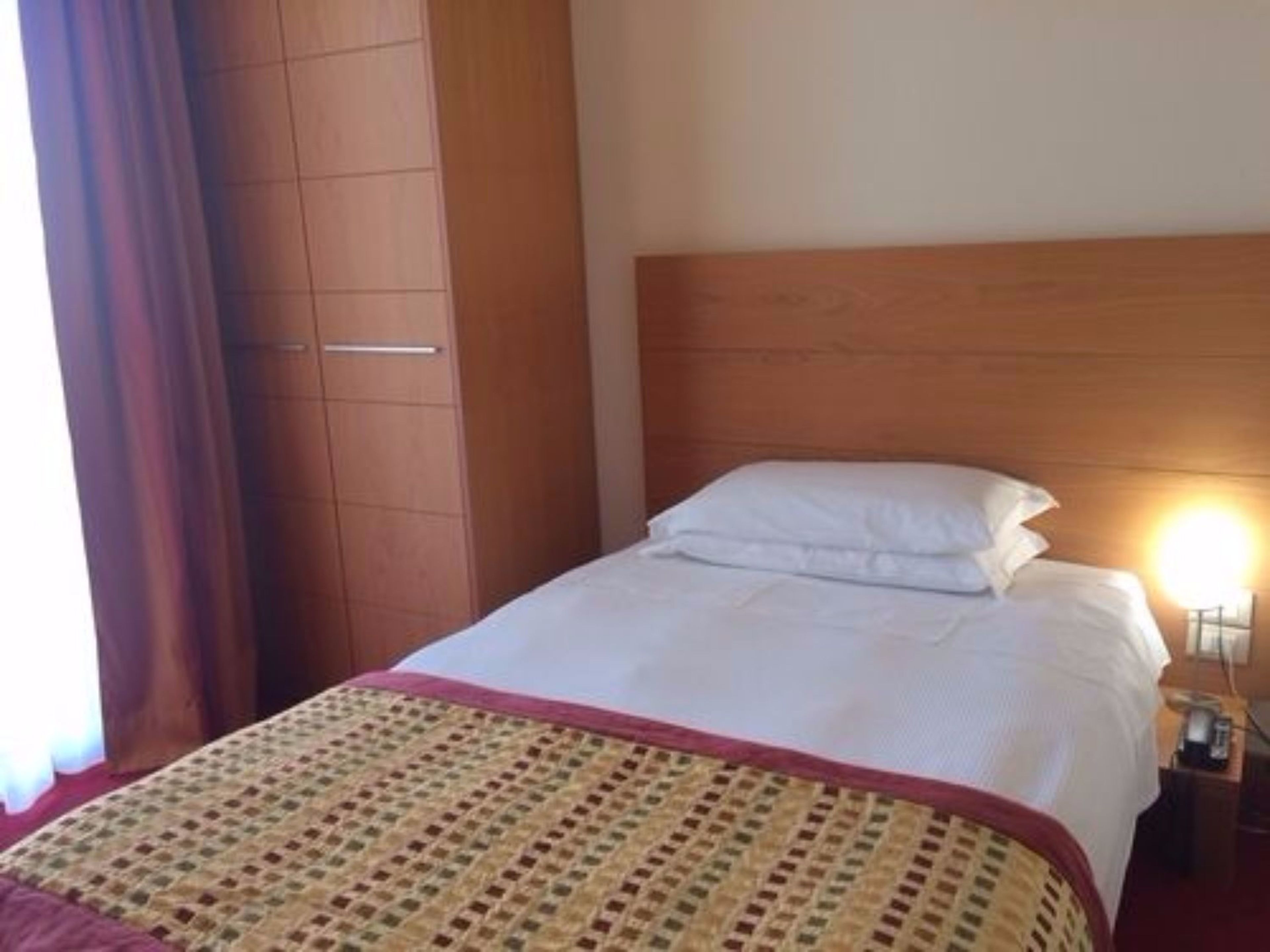 Una habitación de hotel con cama, cabecera de madera, mesita de noche con lámpara y armario.