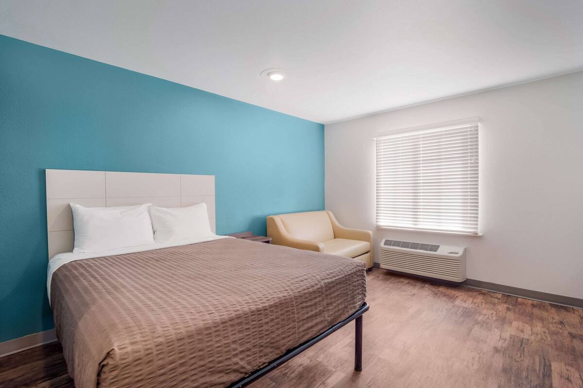 Een hotelkamer met een bed, een stoel, een raam met jaloezieën en een plafondlamp.