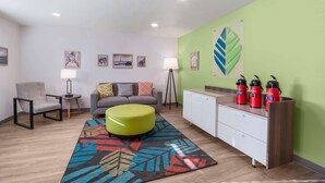 Een moderne woonkamer met een bank, voetenbankje en een kleurrijk tapijt. Er hangen ingelijste foto’s aan de muur, een lamp en een kast met rode thermosflessen.