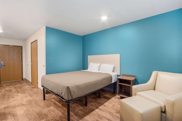 A bedroom with a bed, a nightstand, a chair, and a wooden floor. - WoodSpring Suites North Charleston Airport I-526 (North Charleston)