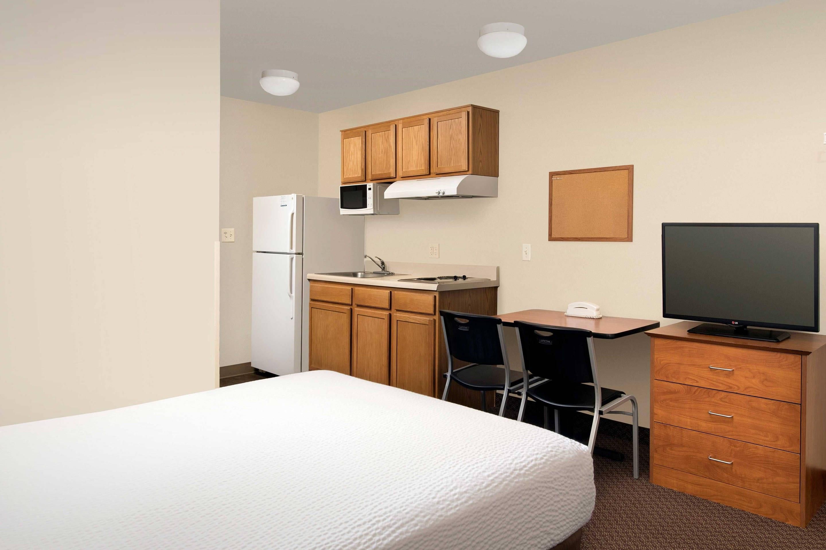 Habitación de hotel compacta con cama, kitchenette, escritorio, silla y televisor.