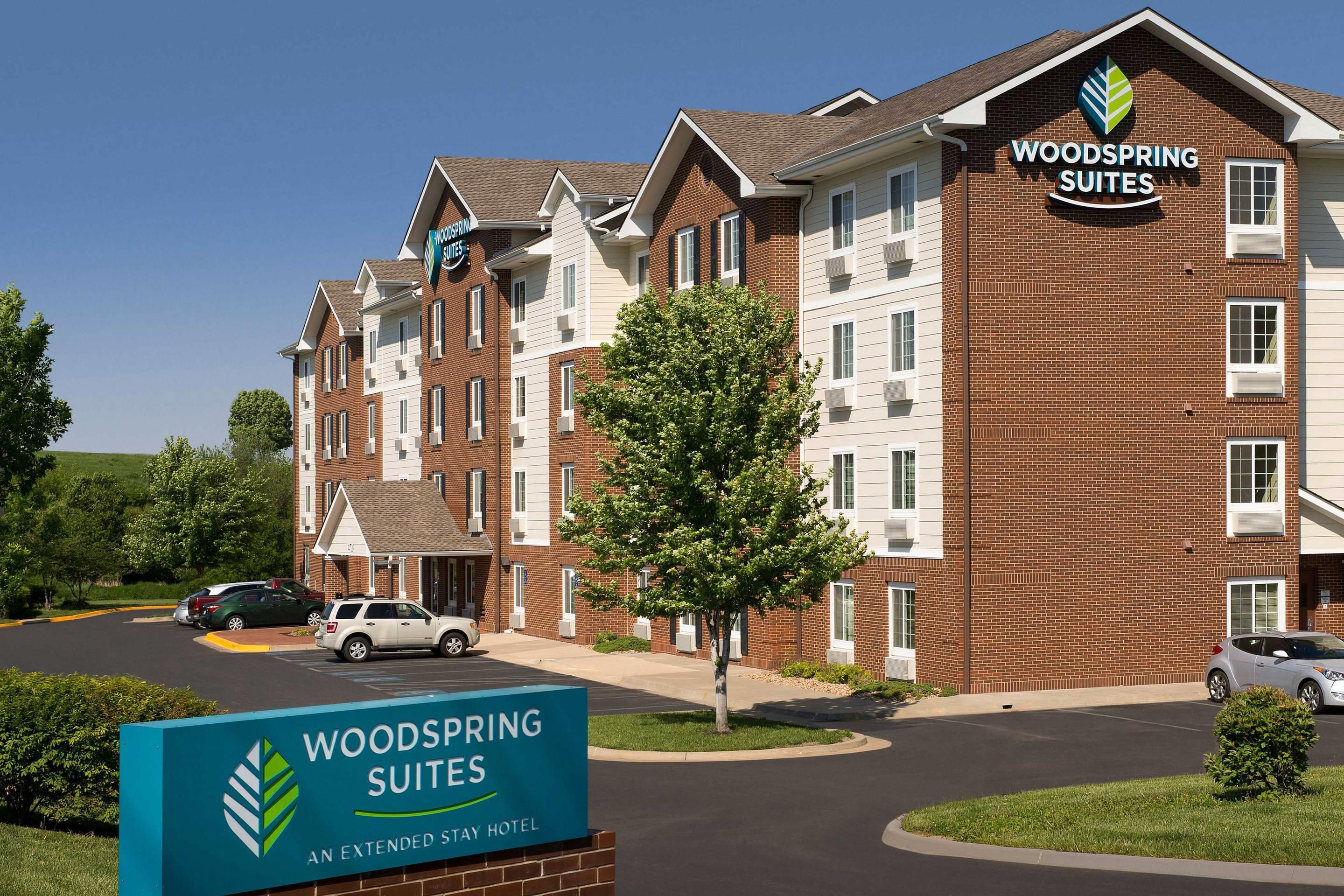 Un hotel de varios pisos con fachada de ladrillo, un cartel que indica que es un Woodspring Suites Extended Stay Hotel, y un estacionamiento con automóviles.