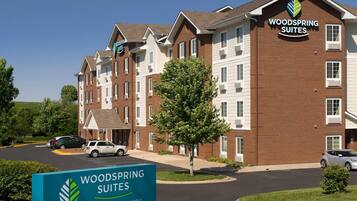 Un hotel de varios pisos con fachada de ladrillo, un cartel que indica que es un Woodspring Suites Extended Stay Hotel, y un estacionamiento con automóviles.