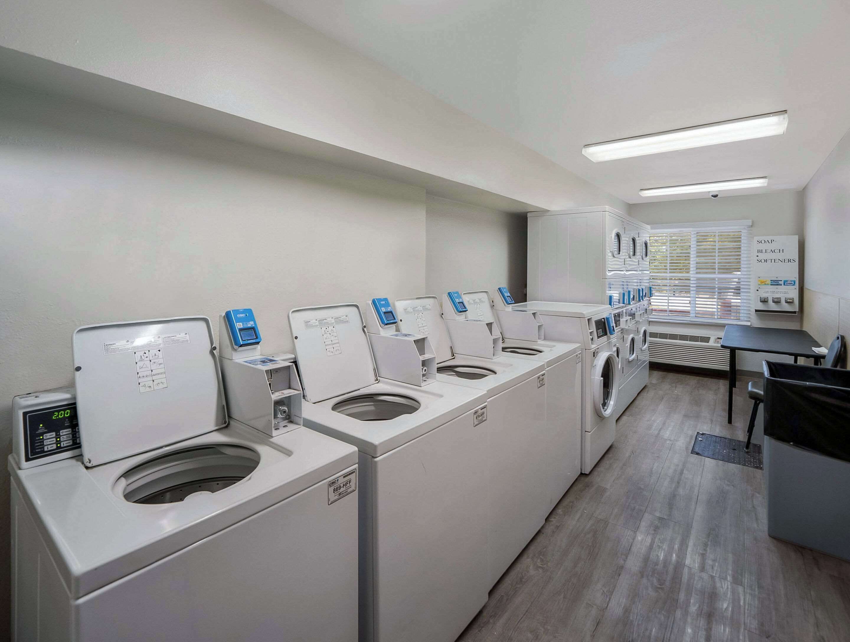 A laundry facility with multiple washing machines and dryers, a control panel, and a sign listing available detergents.