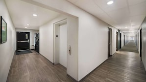 Un pasillo largo y estrecho con piso de madera y paredes blancas. Hay varias puertas a lo largo del pasillo, algunas con letreros encima.
