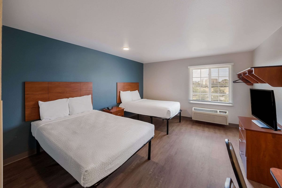 A hotel room with two beds, a desk, a TV, and a window with blinds.