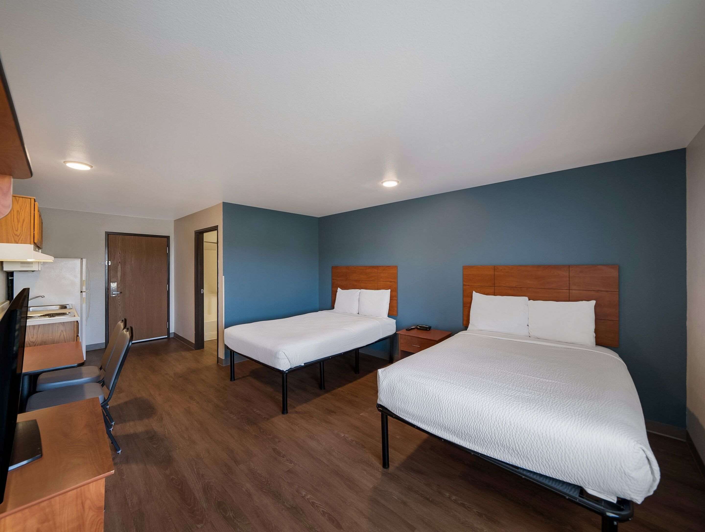 A hotel room with two single beds, a desk, a chair, a small table, and a door leading to a bathroom.