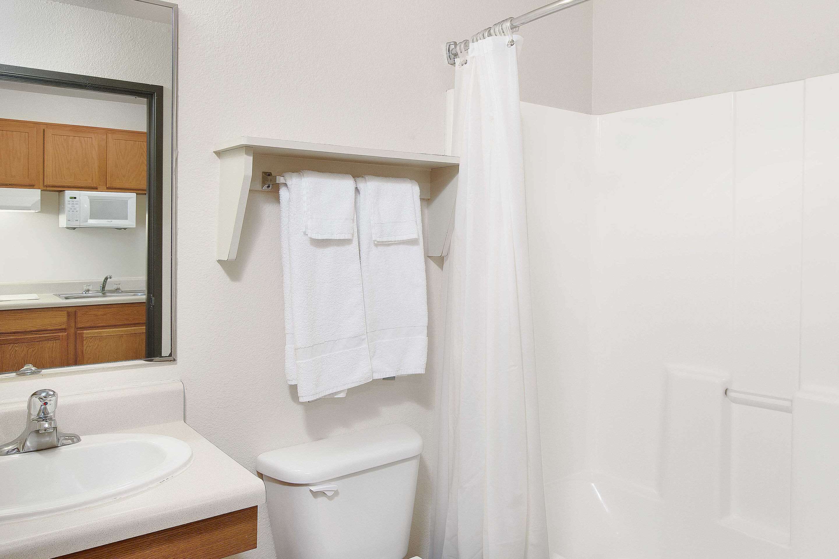 Un baño con lavamanos, inodoro y cortina de ducha blancos. Hay un espejo sobre el lavamanos y un perchero sobre el inodoro.