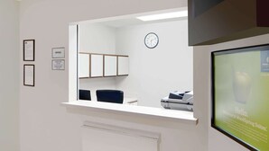 Un área de recepción con un mostrador blanco, un reloj en la pared, diplomas enmarcados y un monitor mostrando un mensaje de bienvenida.