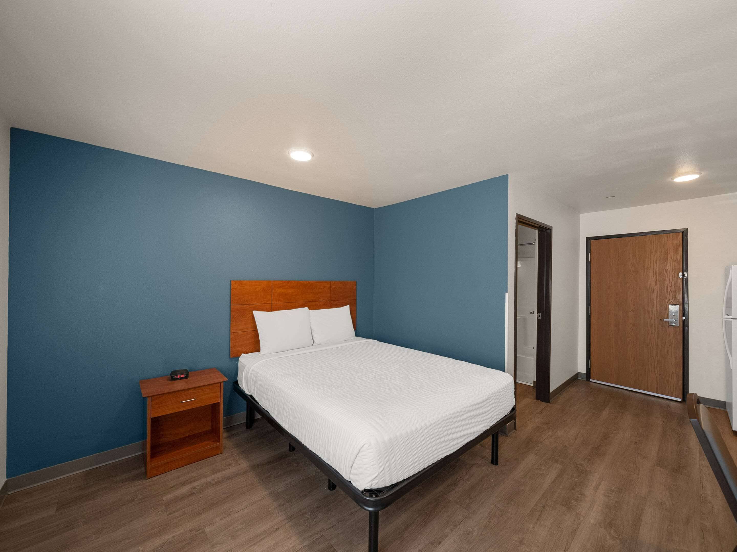 Quarto com cabeceira de madeira, cobreleira branca, mesa de cabeceira e porta à direita.