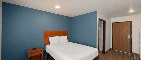 Quarto com cabeceira de madeira, cobreleira branca, mesa de cabeceira e porta à direita.