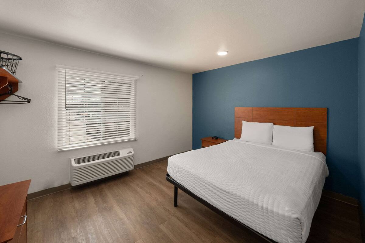 Ein Hotelzimmer mit einem Bett, einem Nachttisch, einem Fenster mit Jalousien, einem Wandheizgerät und einer Holzkommode.