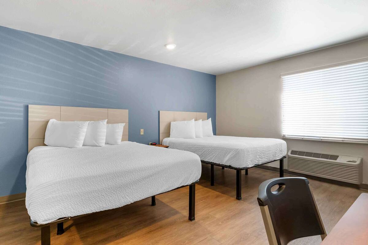 Habitación de hotel con dos camas individuales, suelo de madera, ventana con persianas y aire acondicionado de pared.