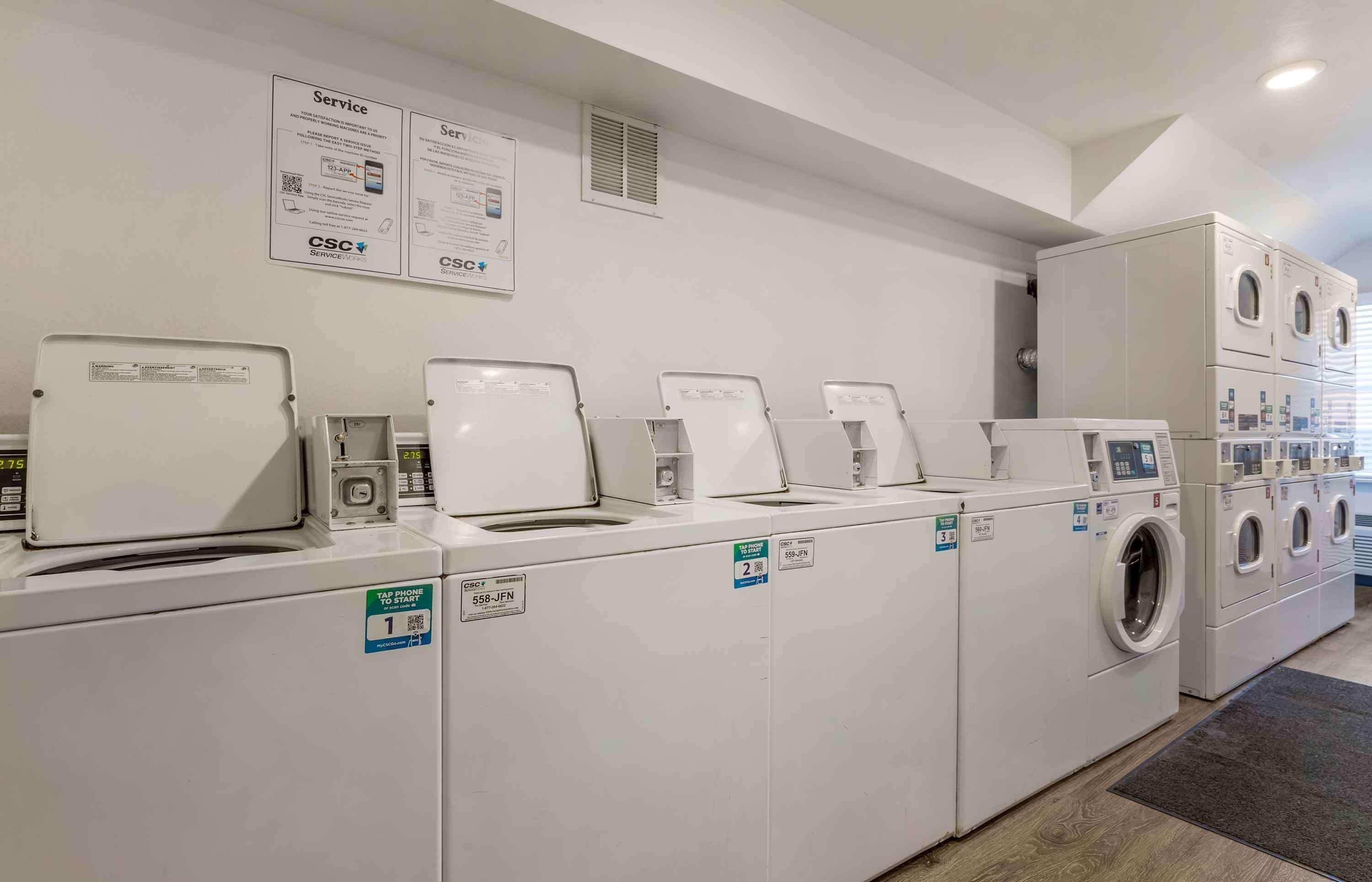 A laundry facility with multiple washing machines and dryers, service information signs, and a QR code for service inquiries.