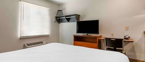 Un dormitorio con una cama grande, un televisor sobre un mueble de madera, un escritorio con una silla y una ventana con persianas.