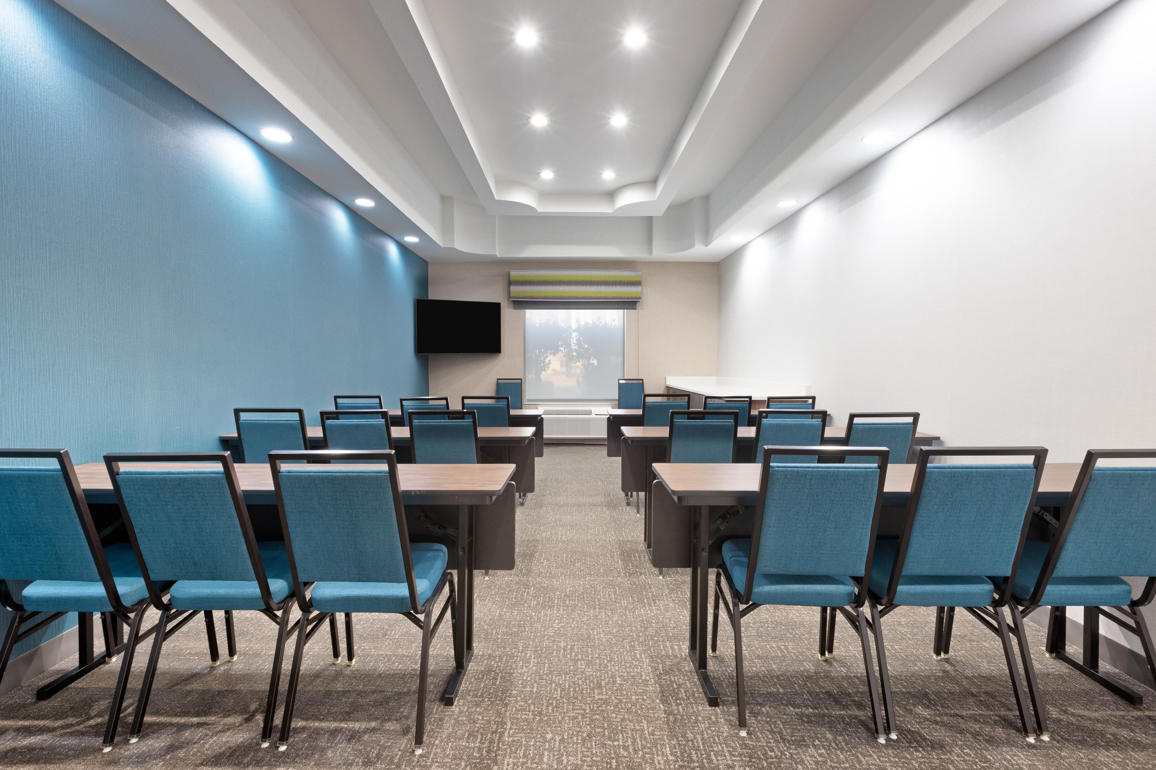 Une salle de conférence avec des rangées de tables et de chaises, un téléviseur à écran plat et une grande fenêtre donnant sur un paysage.