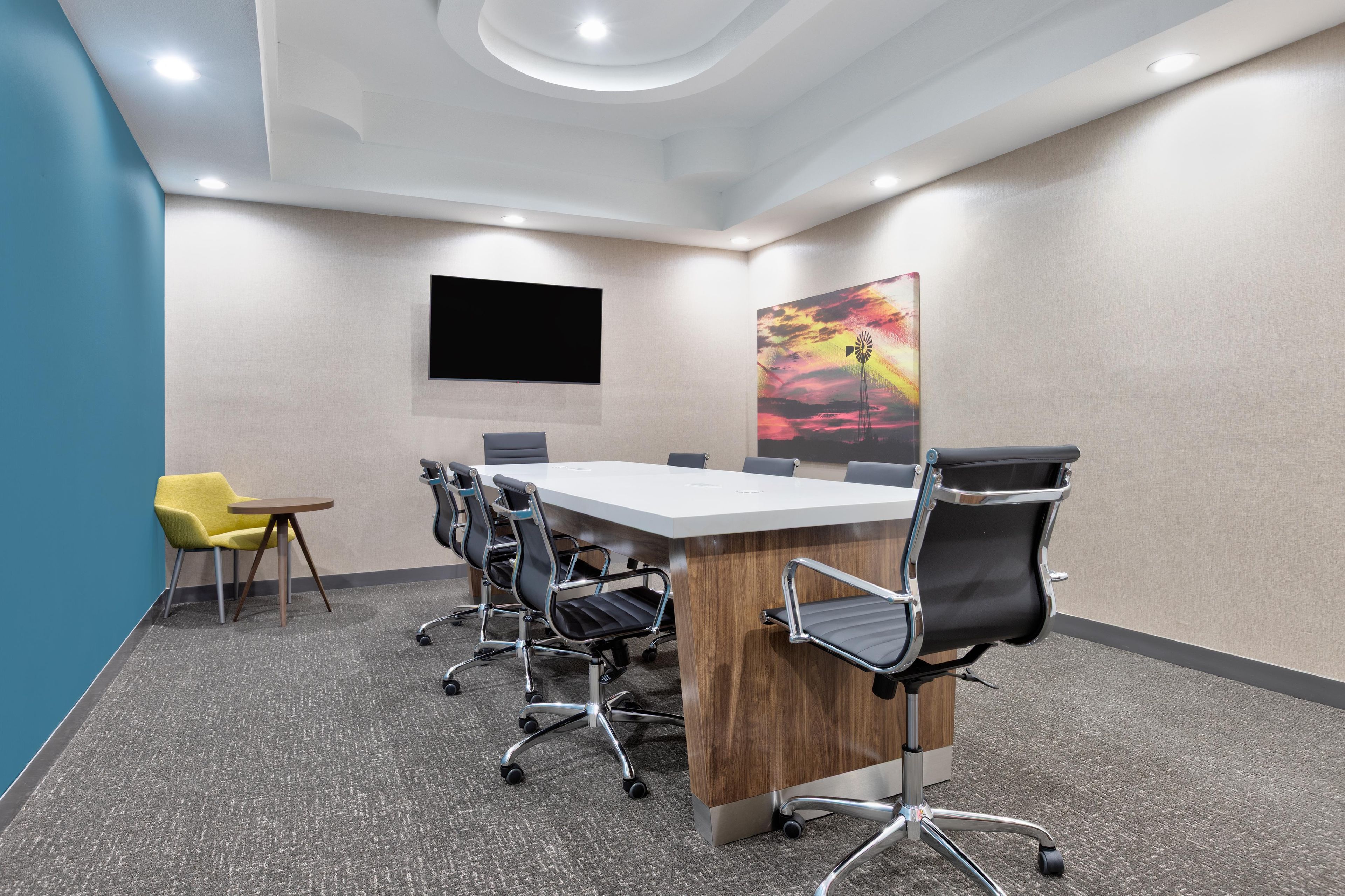A conference room with a long table, six chairs, a TV, and a colorful painting on the wall.