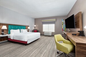 Habitación de hotel con cama, mesitas de noche, escritorio, silla, televisor y ventana con persianas.