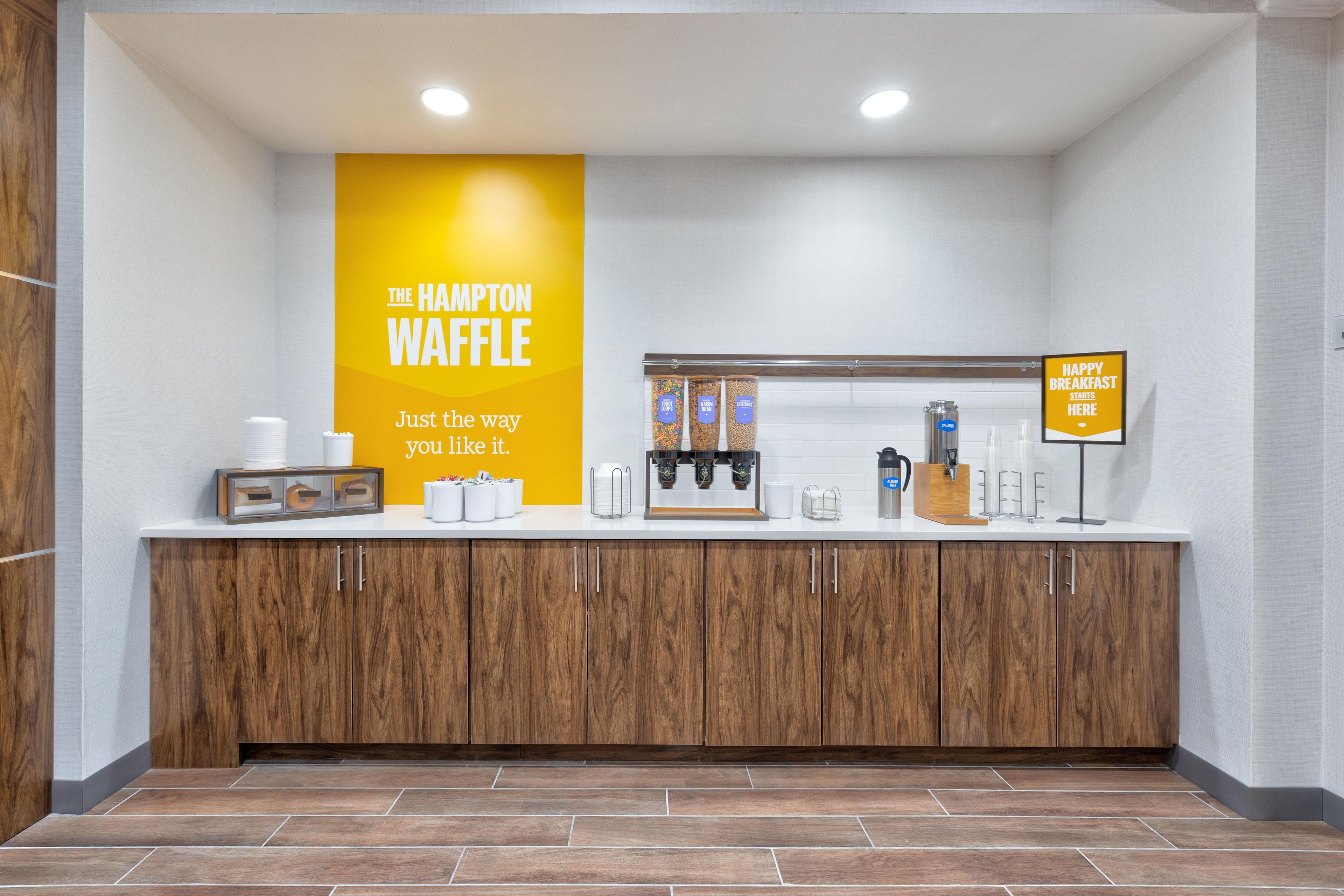 A kitchenette area with a waffle station, coffee machines, and a sign indicating breakfast service.