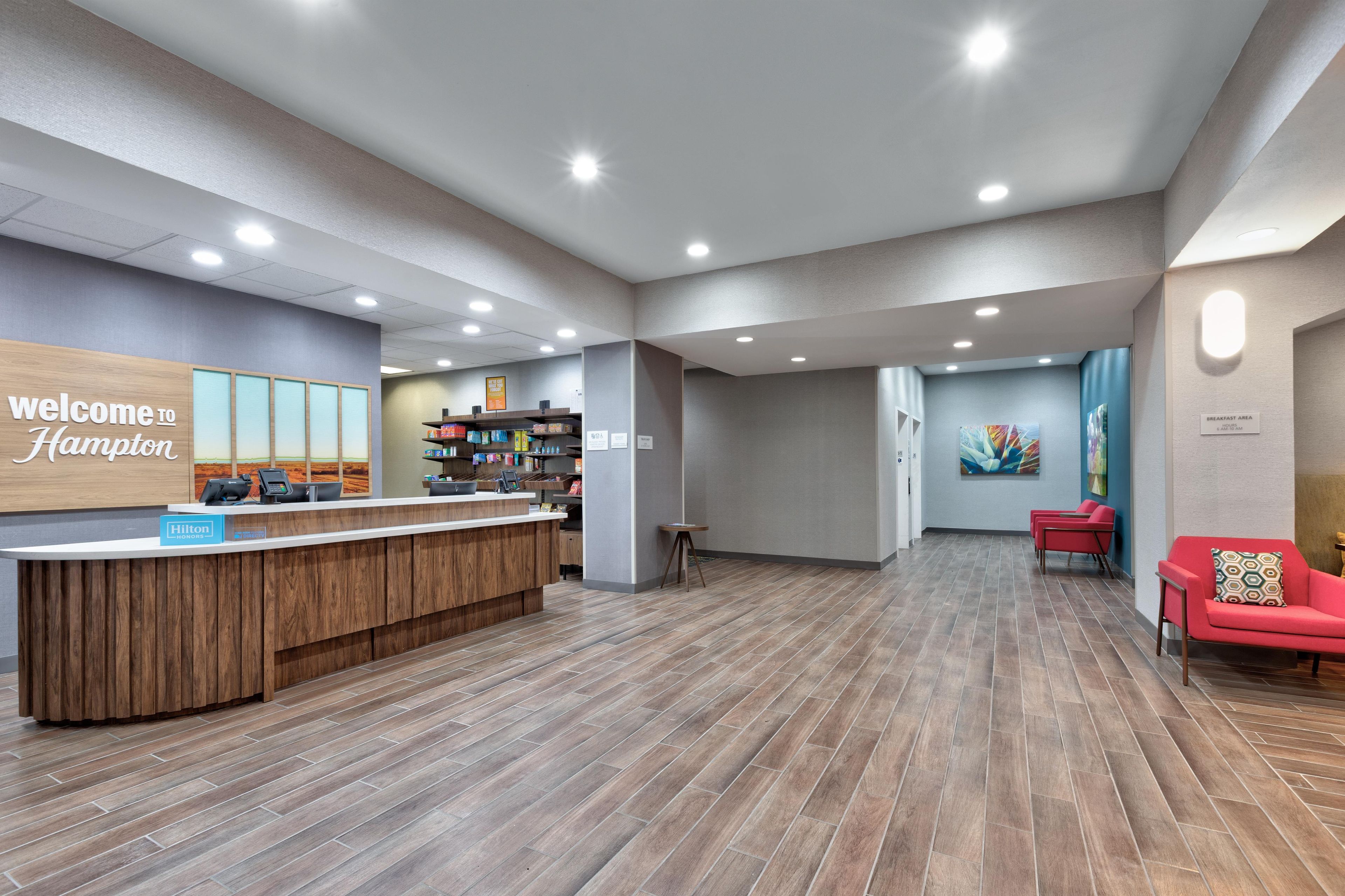 A hotel reception area with a wooden reception desk, a 'Welcome to Hampton' sign, a breakfast area sign, and a seating area with red chairs.