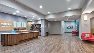 A hotel reception area with a wooden reception desk, a 'Welcome to Hampton' sign, a breakfast area sign, and a seating area with red chairs.