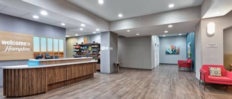 Zona de recepción de hotel con un mostrador de madera, un letrero que dice 'Welcome to Hampton', señalización para el área de desayuno y un espacio de estar con sillas rojas.