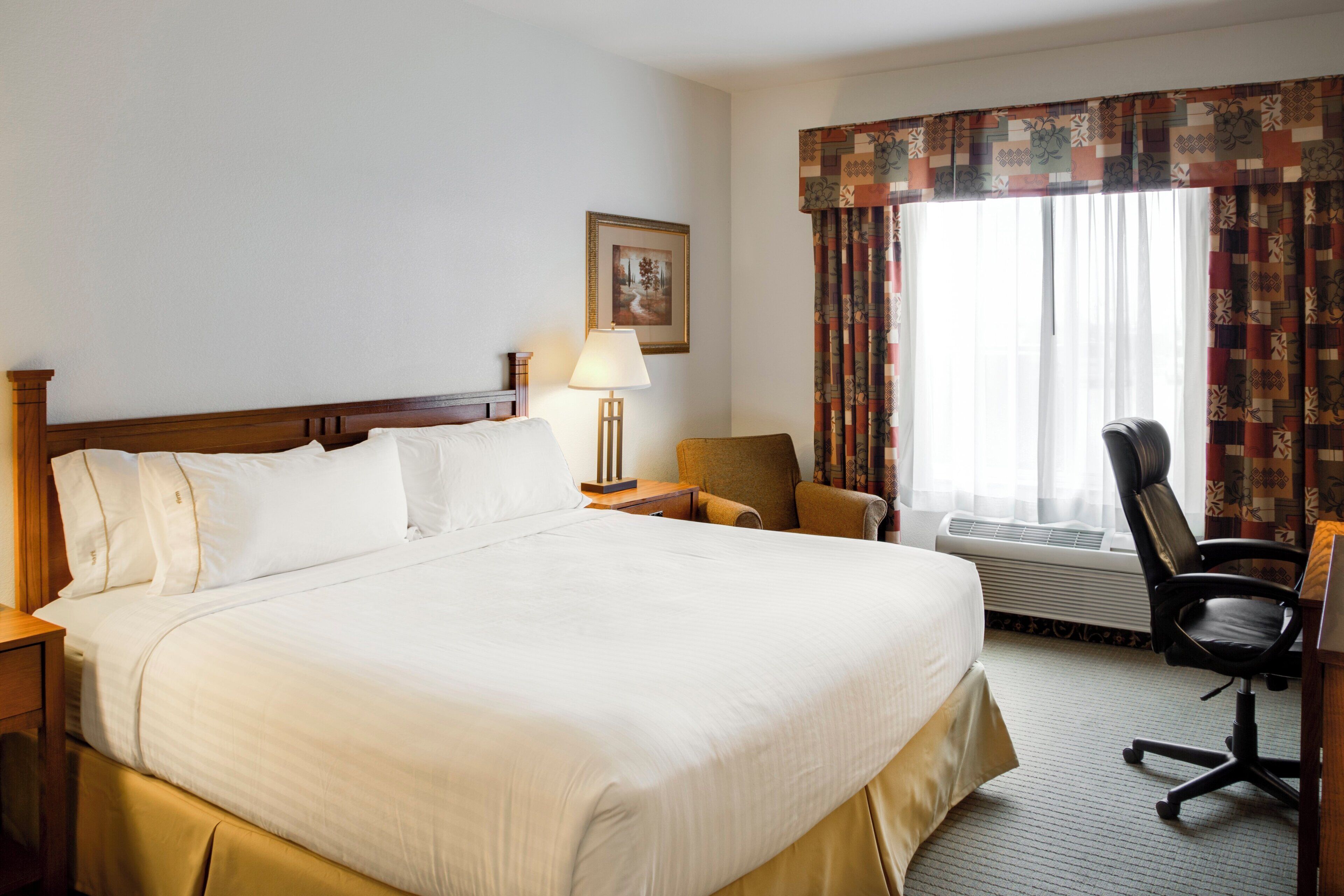 A hotel room with a large bed, a desk with a lamp, a chair, a window with curtains, and a picture on the wall.
