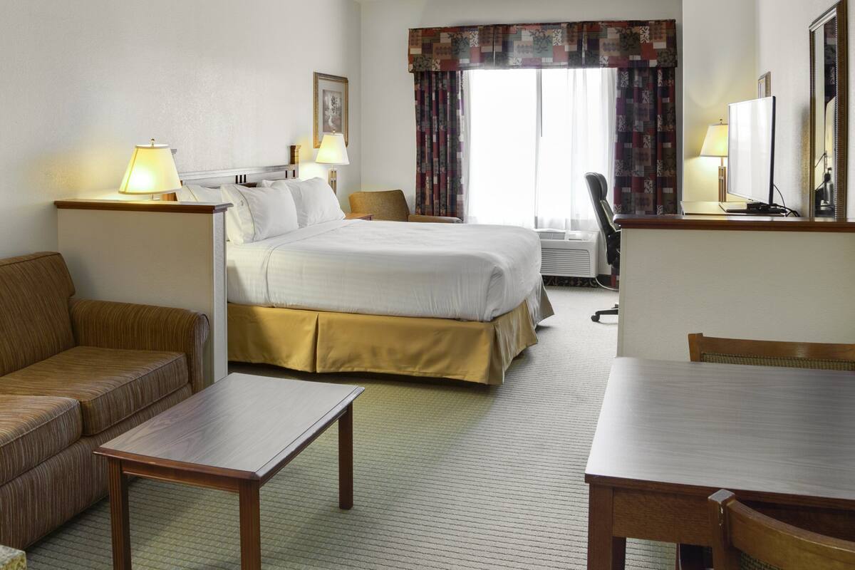 A hotel room with a bed, a sofa, a table, a chair, a desk, a lamp, a mirror, and a window with curtains.