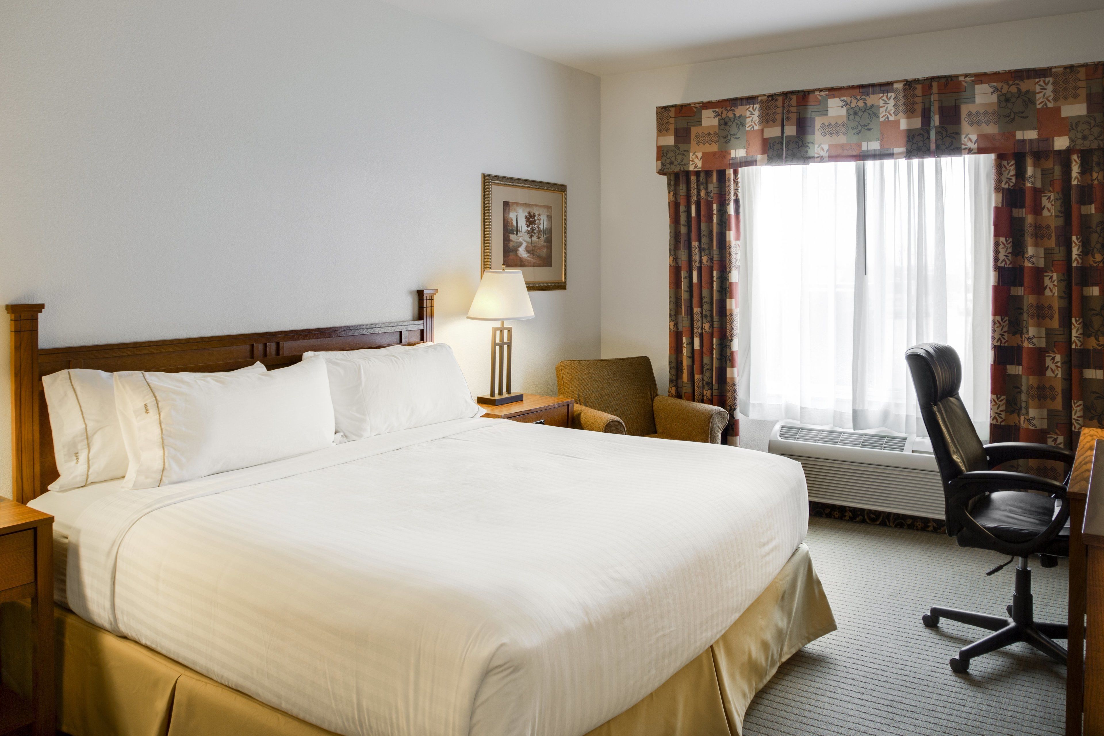 A hotel room with a large bed, a desk, a chair, a lamp, and a window with curtains.