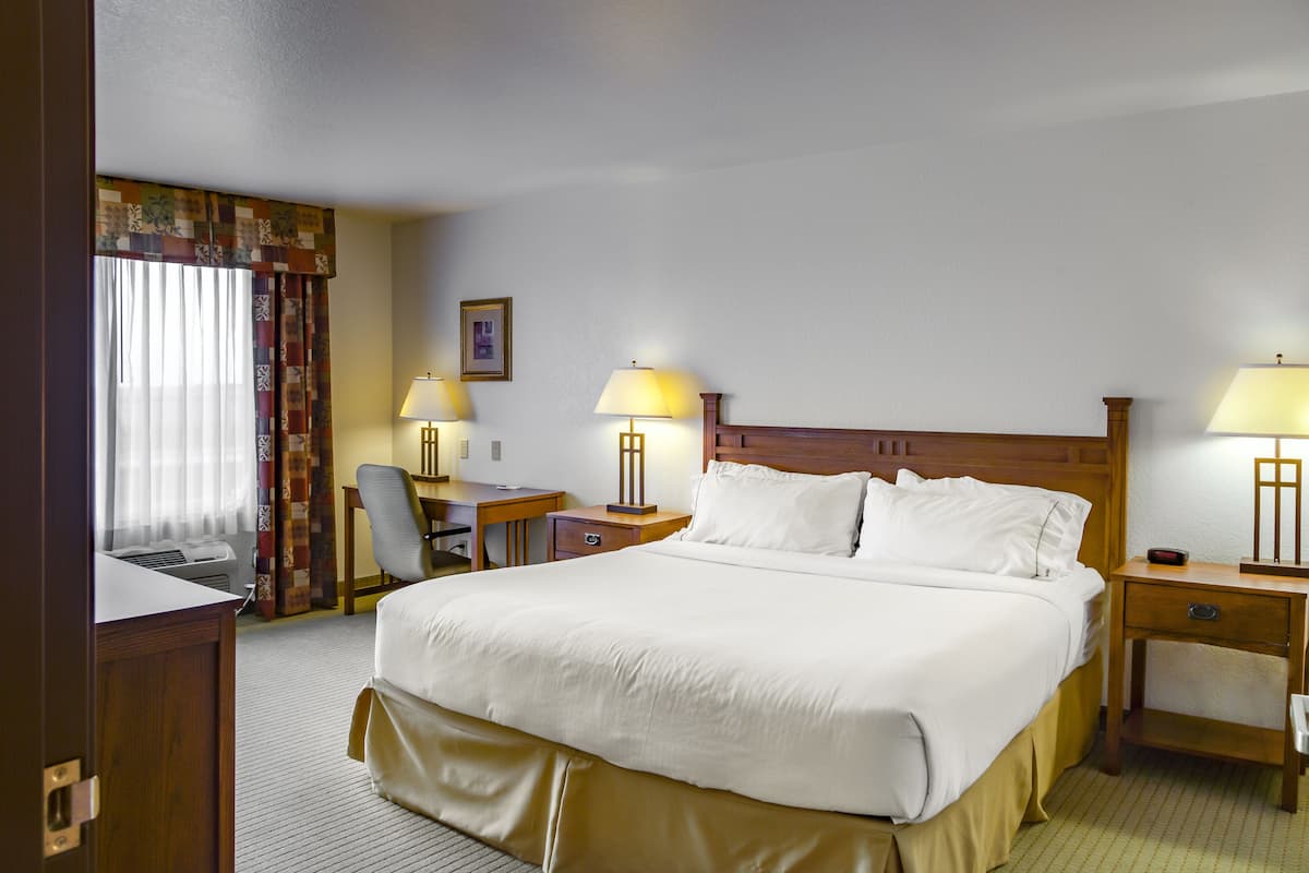 A hotel room with a large bed, two bedside tables with lamps, a desk with a chair, and a window with curtains.