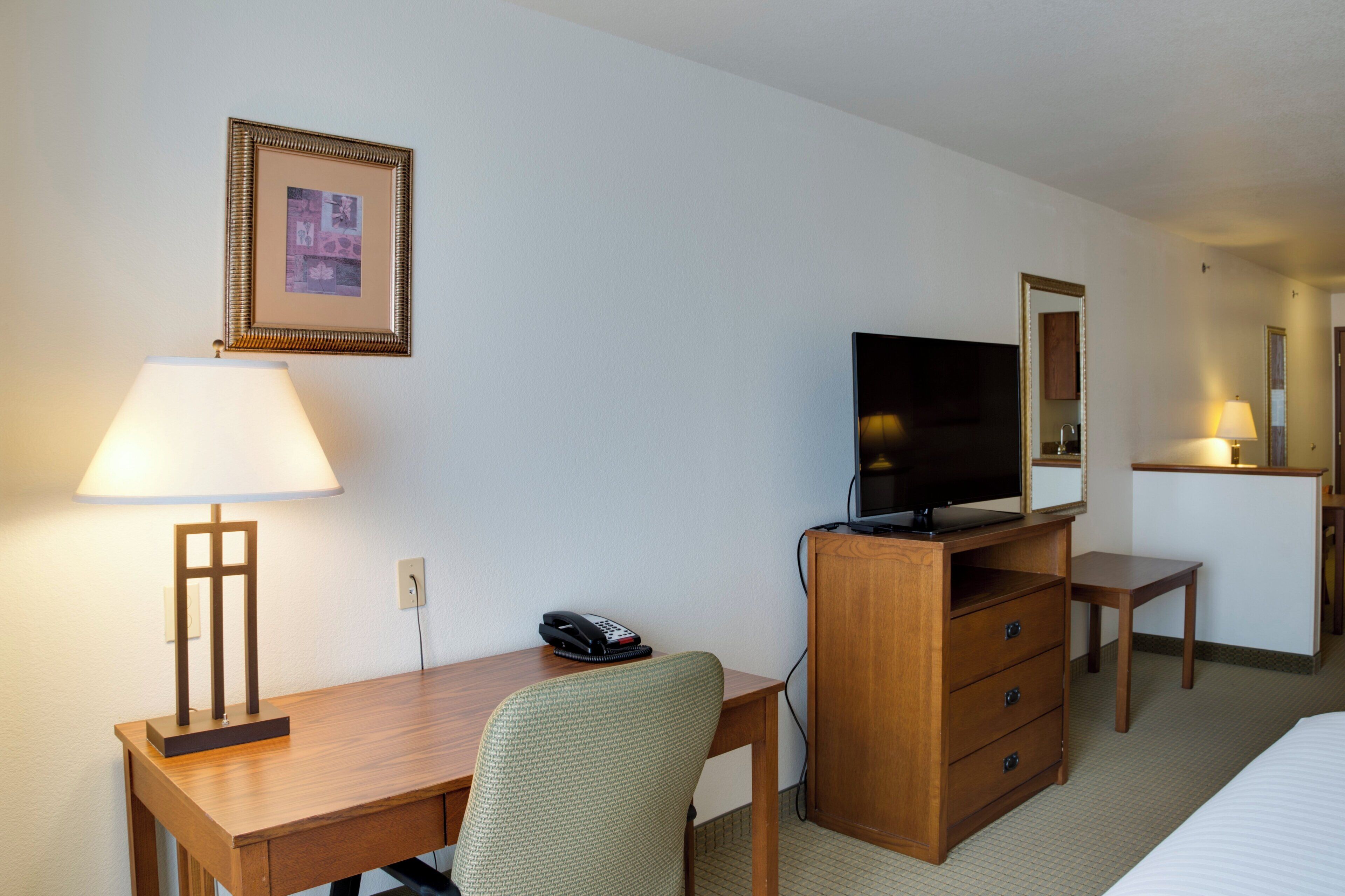 Habitación de hotel con un escritorio de madera, una silla, un televisor sobre un mueble de madera, un teléfono, un cuadro en la pared y un espejo.