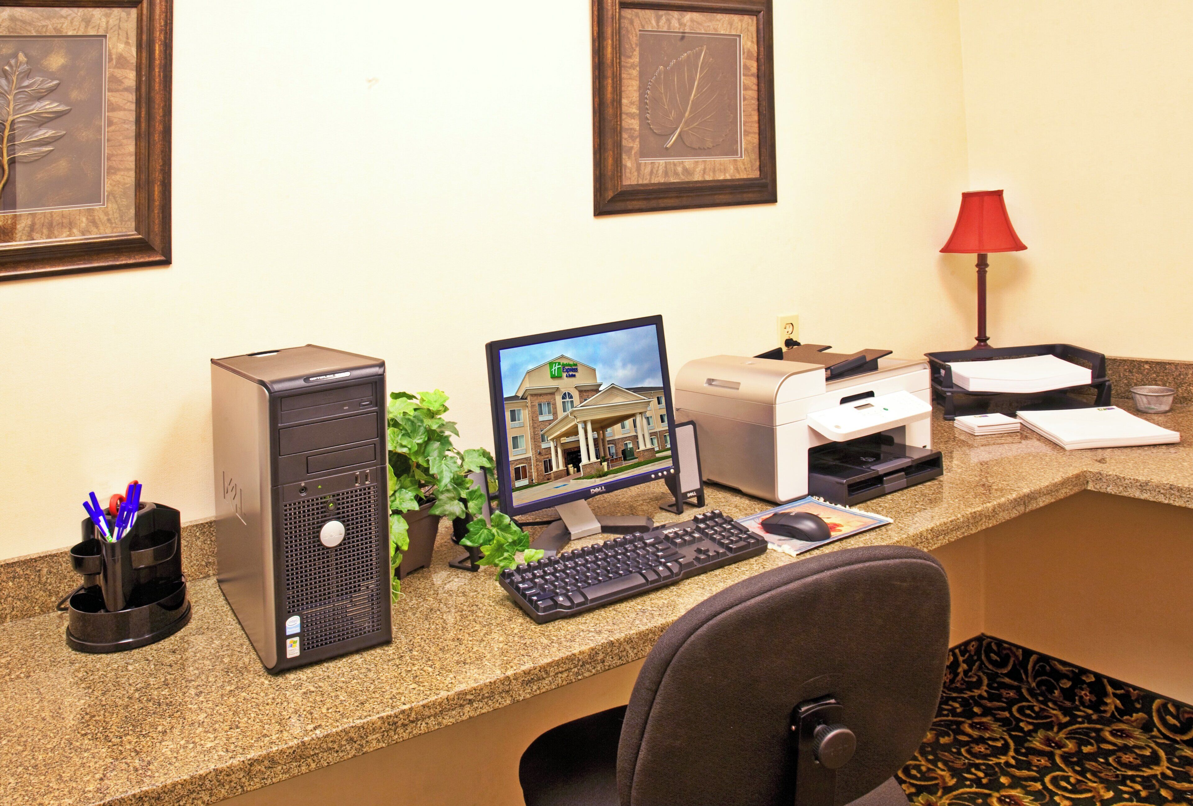 Un mostrador de recepción de hotel con una computadora de escritorio, una impresora, una lámpara de escritorio y un cuadro en la pared.