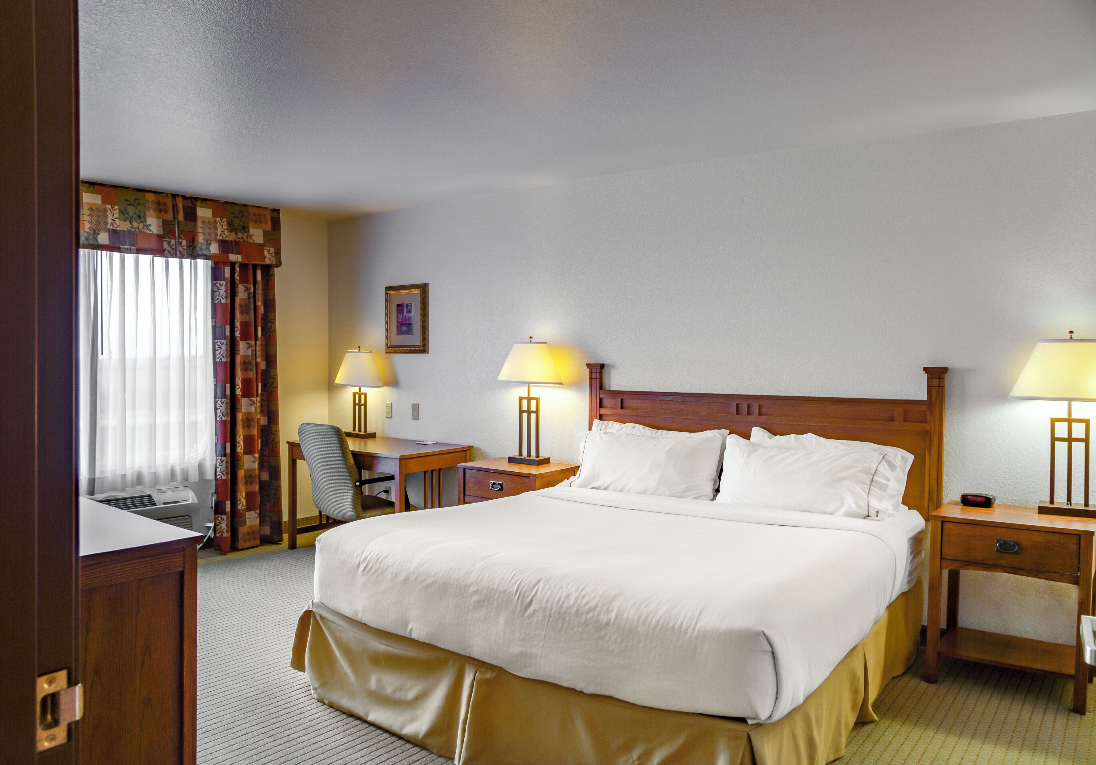 Quarto de hotel com uma cama grande, duas mesas de cabeceira com luminárias, uma escrivaninha, uma cadeira e uma janela com cortinas.