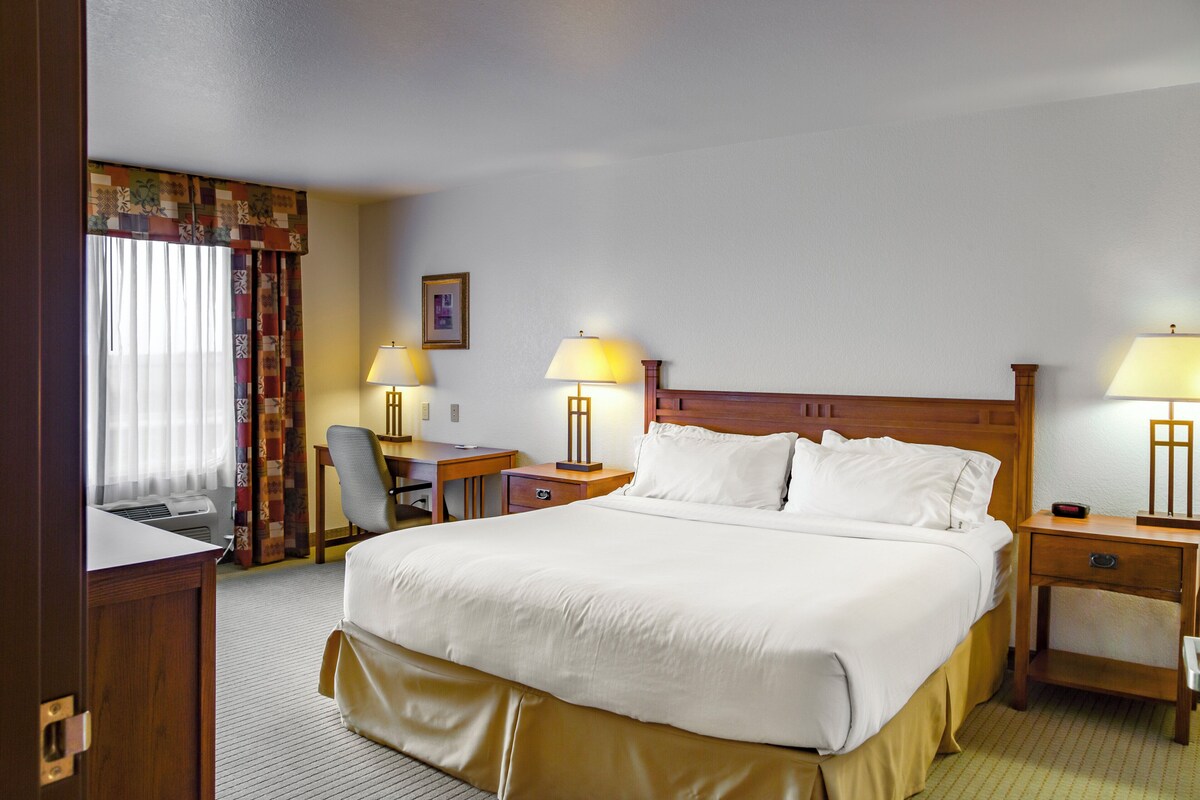 Habitación de hotel con una cama grande, dos mesitas de noche con lámparas, un escritorio, una silla y un ventanal con cortinas.