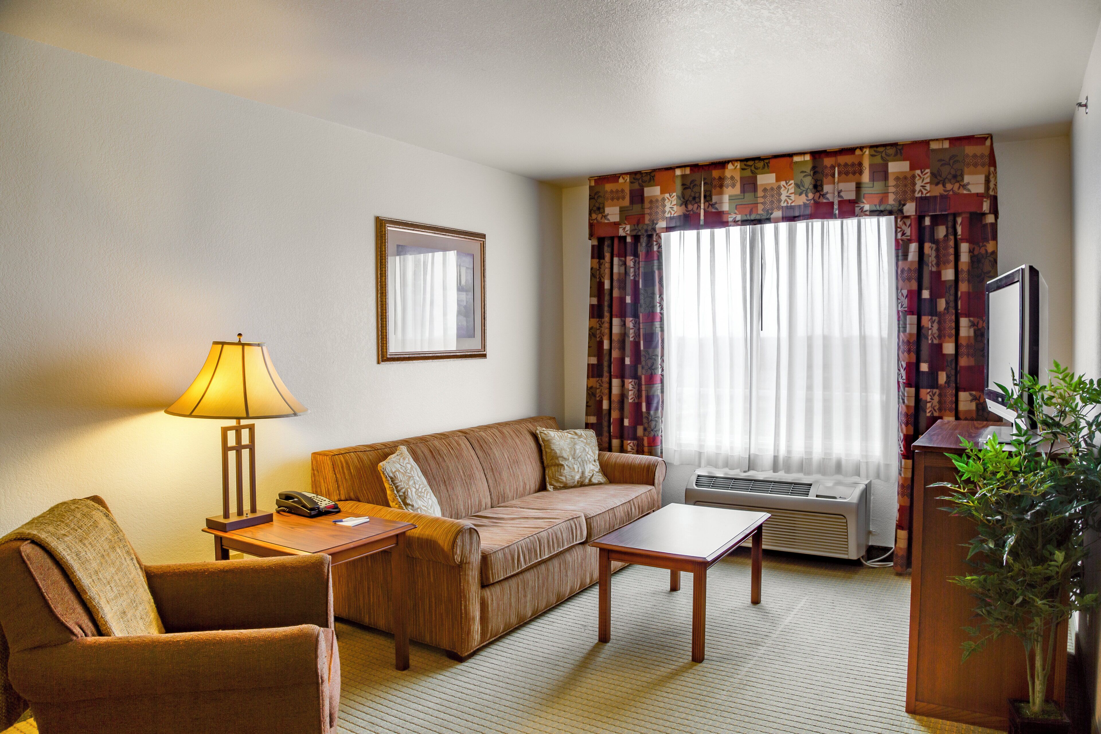 A hotel room with a sofa, two armchairs, a small table, a lamp, a mirror, an air conditioner, and a television.